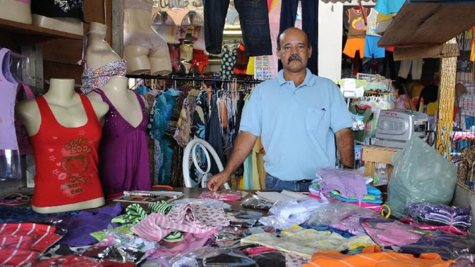 Comerciante atrás do balcão de uma loja de roupas de pequeno porte, com peças expostas ao redor.
