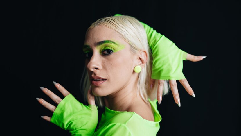 A cantora Juliana Linhares, de cabelo platinado e roupa verde-limão, com maquiagem verde nos olhos, em foto de estúdio sobre fundo escuro