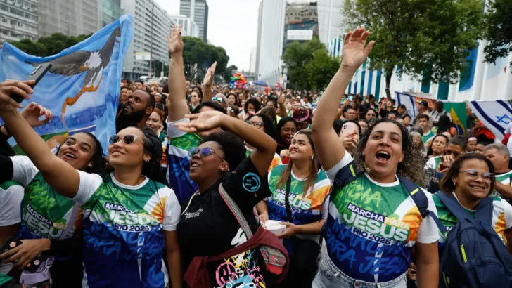 Multidão de fiéis com camisetas da Marcha para Jesus canta com os braços erguidos em avenida