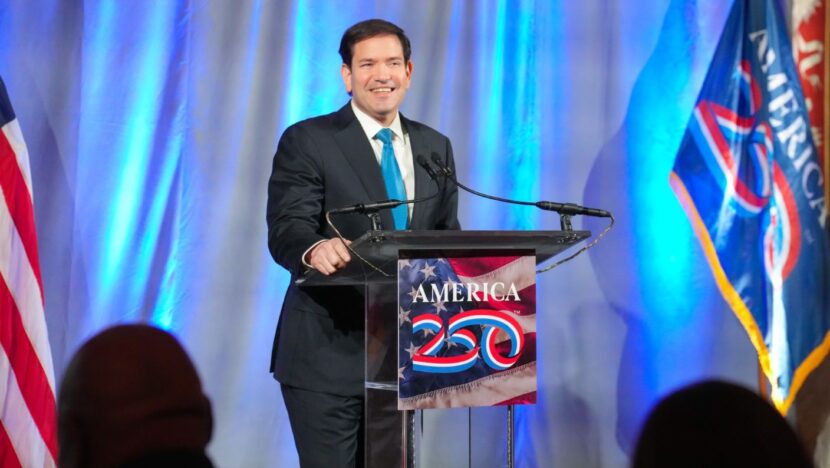 Marco Rubio sorri em púlpito com a inscrição America 250, diante de bandeiras dos Estados Unidos