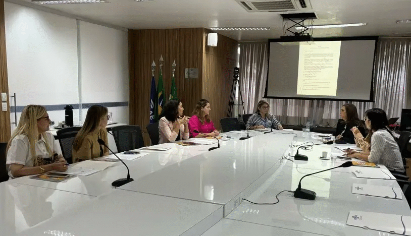 Mulheres sentadas em mesa de reunião com microfones acompanham apresentação em telão
