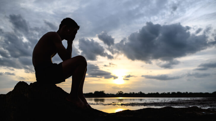um homem sentado dobrou os joelhos segurando as maos no rosto na base da arvore e havia agua ao redor