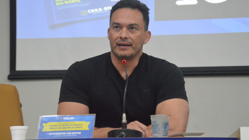 Styvenson Valentim, de camisa preta, sentado à mesa diante de um microfone