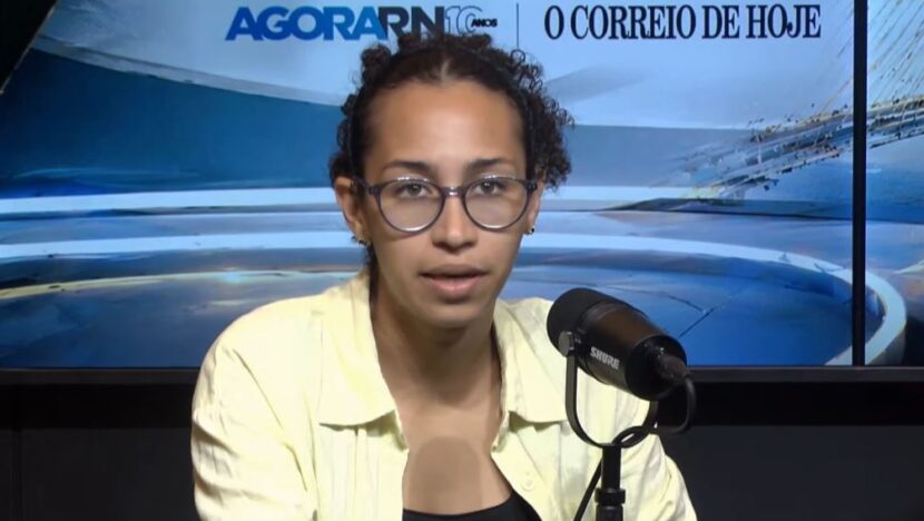 Anne Lagartixa, de camisa clara e óculos, fala ao microfone em estúdio de televisão com o painel do Agora RN e de O Correio de Hoje ao fundo