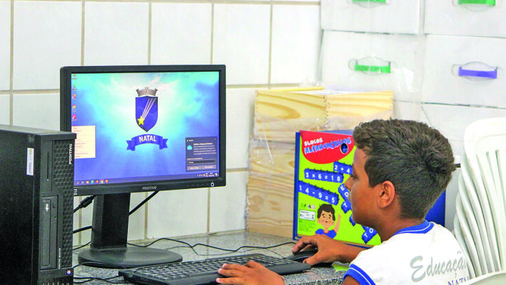Estudante sentado diante de um computador em ambiente escolar, com livros ao lado do monitor