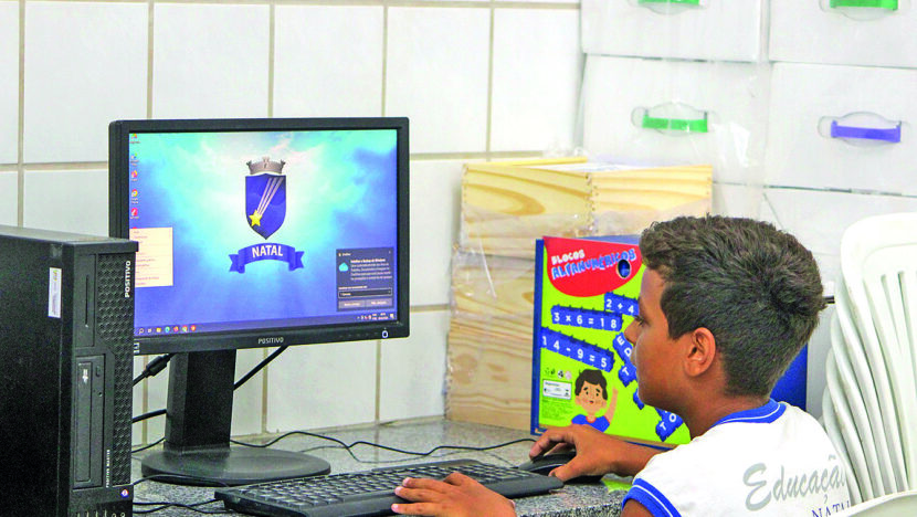 Estudante sentado diante de um computador em ambiente escolar, com livros ao lado do monitor