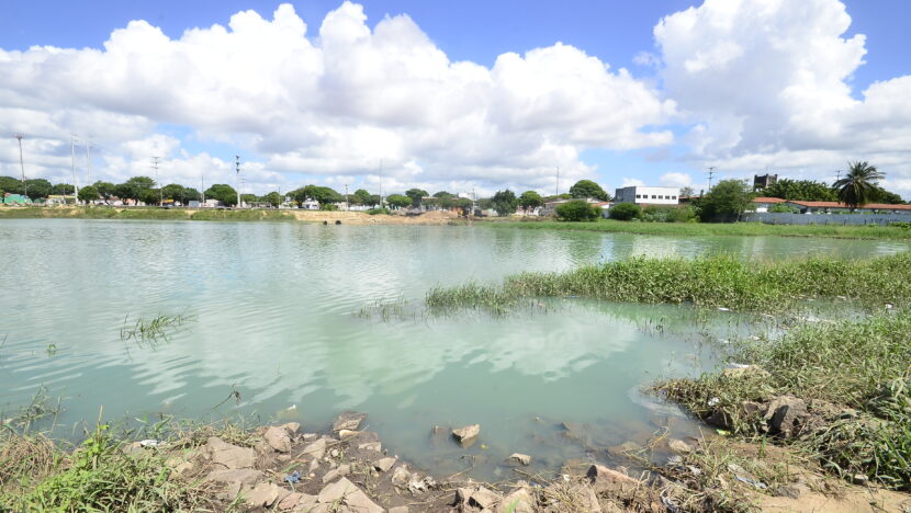 Lagoa de captação cheia de água esverdeada, com vegetação e entulho na margem em primeiro plano e casas ao fundo, sob céu com nuvens