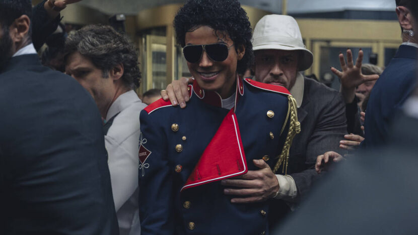 Ator caracterizado como Michael Jackson, com uniforme azul e vermelho e óculos escuros, cercado por pessoas