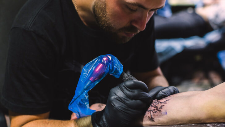 Profissional realiza tatuagem no braço de uma pessoa utilizando equipamento e luvas de proteção