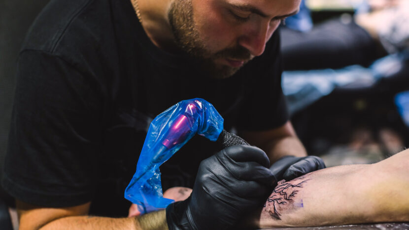 Profissional realiza tatuagem no braço de uma pessoa utilizando equipamento e luvas de proteção