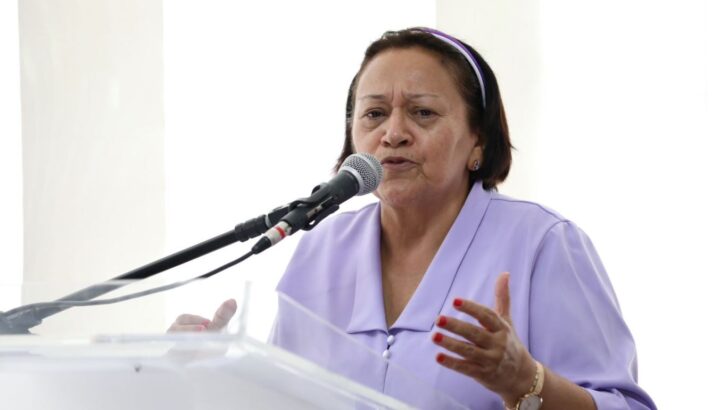 Mulher de roupa lilás fala ao microfone durante evento