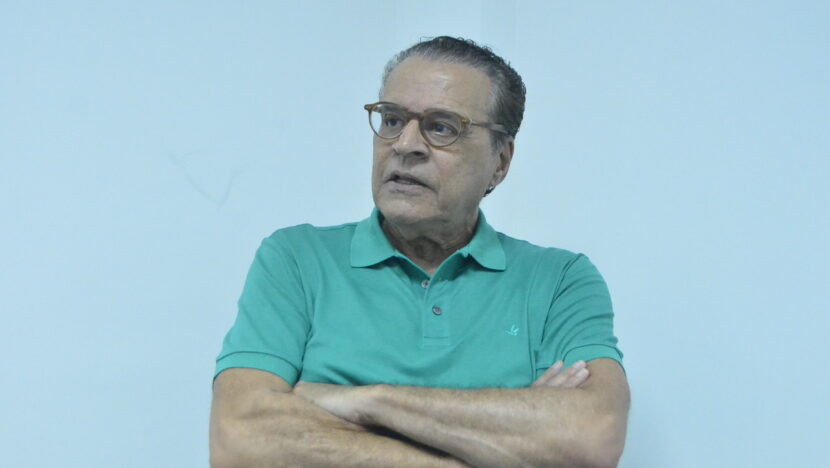 Homem de óculos e camisa polo verde aparece sentado e gesticula com as mãos durante uma conversa
