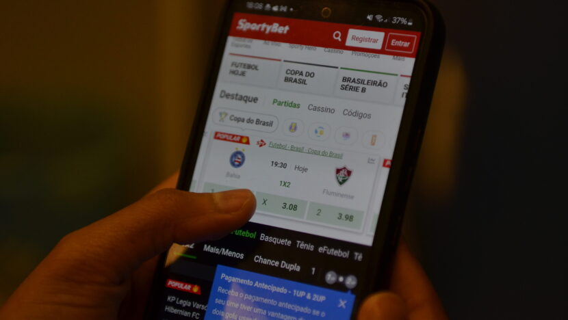 Mão segura celular que mostra eventos esportivos e jogos virtuais on-line - Foto: José Aldenir