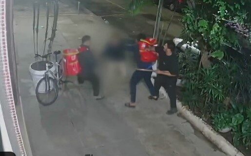 Imagem de câmera de segurança mostra homens durante agressão a uma pessoa em uma calçada, com uma bicicleta ao lado.