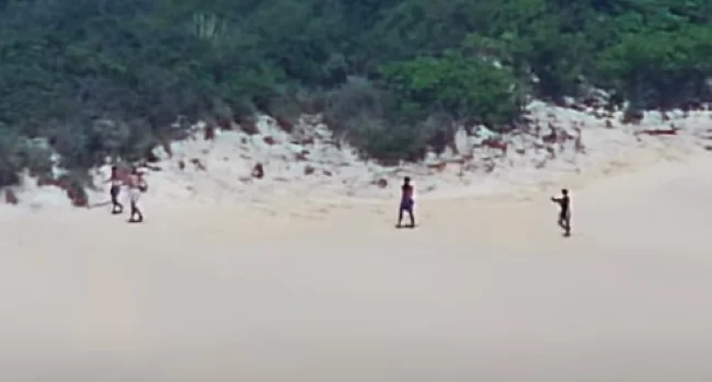 Quatro pessoas caminham pela faixa de areia próxima à vegetação do Morro do Careca