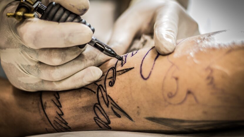 Profissional usando luvas realiza tatuagem no braço de uma pessoa com máquina própria para o procedimento