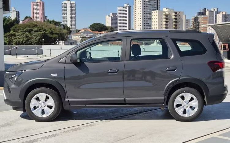Chevrolet Spin cinza vista de perfil, estacionada em área aberta, com prédios ao fundo