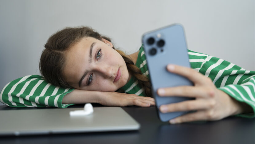 Jovem apoia a cabeça sobre o braço enquanto olha para a tela de um celular sobre uma mesa com um computador portátil