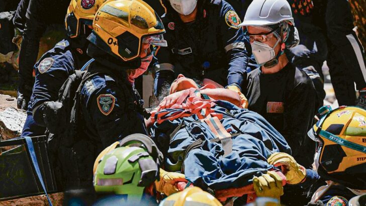 Equipes de emergência com capacetes e equipamentos de proteção transportam uma pessoa em uma maca durante operação de resgate