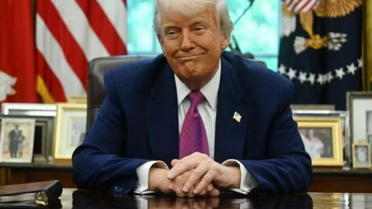 Donald Trump sentado à mesa, com bandeiras dos Estados Unidos e símbolos oficiais ao fundo