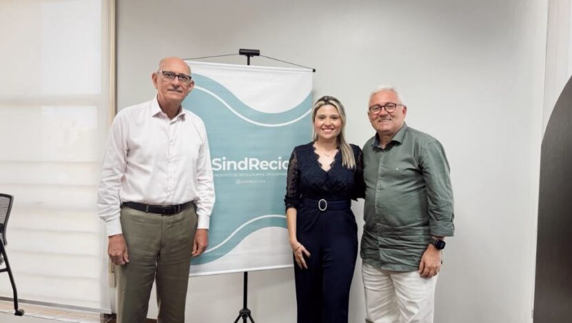 Fernanda Medeiros posa entre dois homens em frente a um banner do Sindrecicla-RN