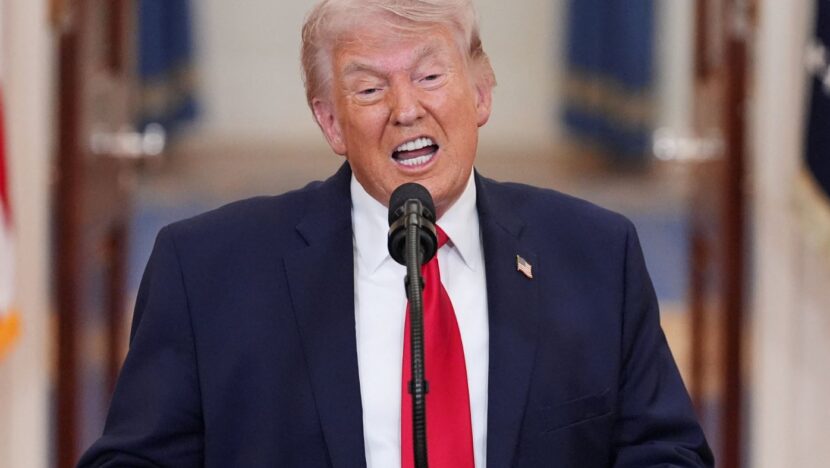 Donald Trump fala ao microfone durante pronunciamento, usando terno escuro e gravata vermelha