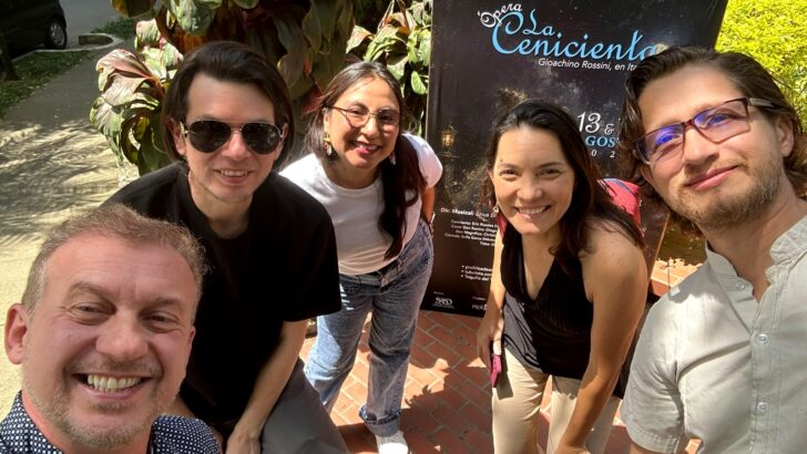 Grupo de cinco pessoas posa para foto diante de cartaz da ópera “La Cenerentola”, em área externa