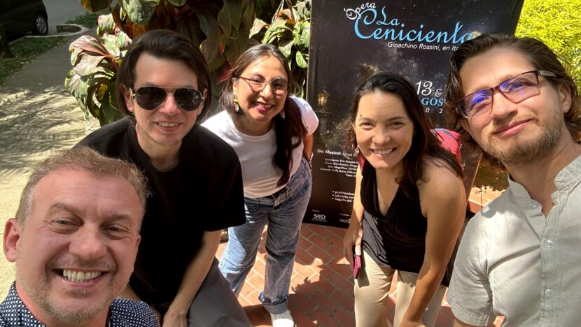 Grupo de cinco pessoas posa para foto diante de cartaz da ópera “La Cenerentola”, em área externa