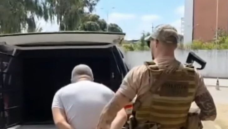 Homem com as mãos para trás é conduzido por agente de segurança em direção a um veículo
