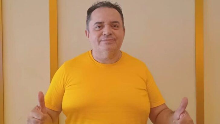 Homem de camiseta amarela posa diante de uma parede e faz sinal de positivo com as duas mãos