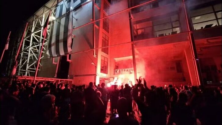 Torcedores reunidos durante a noite em frente ao estádio do PAOK, iluminado por sinalizadores vermelhos