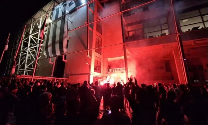 Torcedores reunidos durante a noite em frente ao estádio do PAOK, iluminado por sinalizadores vermelhos