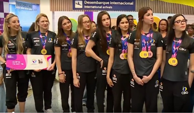 Integrantes da seleção brasileira de ginástica rítmica reunidas no aeroporto, usando uniformes da equipe e medalhas no pescoço