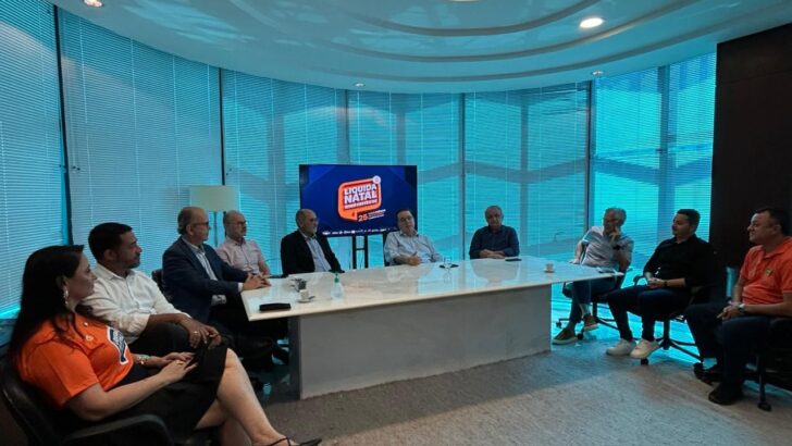 Grupo de pessoas reunido em uma sala, ao redor de uma mesa, com tela exibindo a identidade visual da Liquida Natal e a referência aos 25 anos da campanha