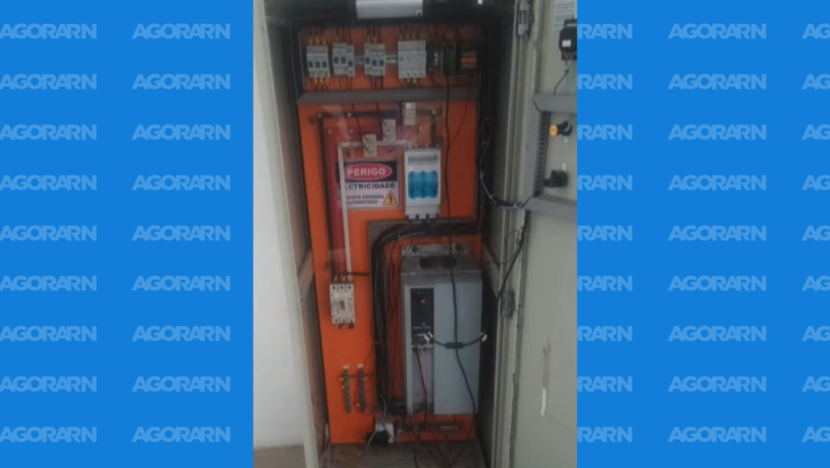 Armário elétrico aberto mostra painel laranja, disjuntores e fiação exposta, além de uma placa de advertência sobre risco de eletricidade