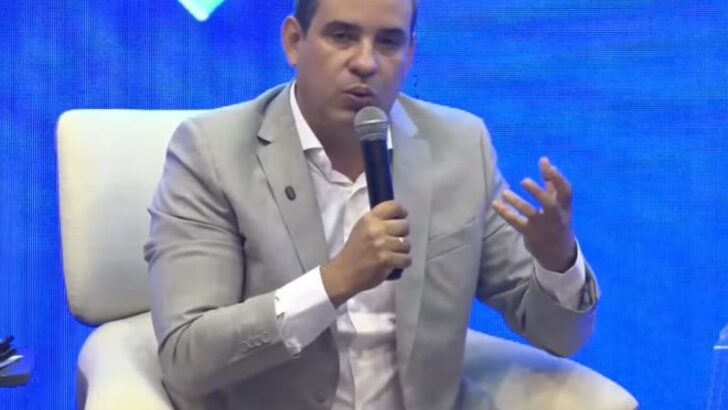 Cadu Xavier, vestido com terno cinza, segura um microfone e gesticula durante a sabatina do Fórum Caminhos do RN