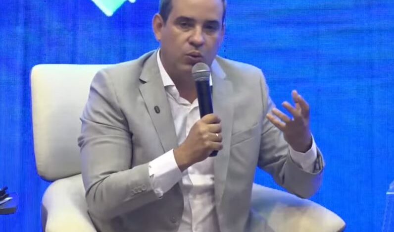 Cadu Xavier, vestido com terno cinza, segura um microfone e gesticula durante a sabatina do Fórum Caminhos do RN