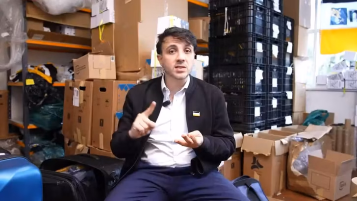 Homem aparece sentado em ambiente com caixas e outros materiais armazenados ao fundo