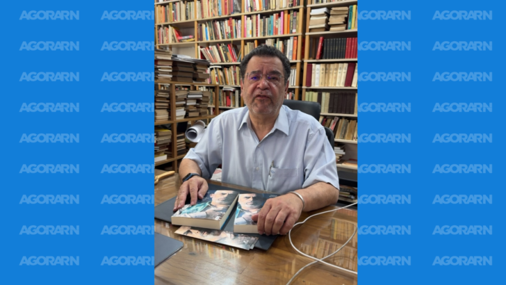 Rui Costa Pimenta aparece sentado diante de uma mesa com livros, em ambiente cercado por estantes com diversas obras