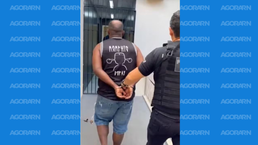 Homem algemado, de costas, é conduzido por policial em corredor de uma unidade.
