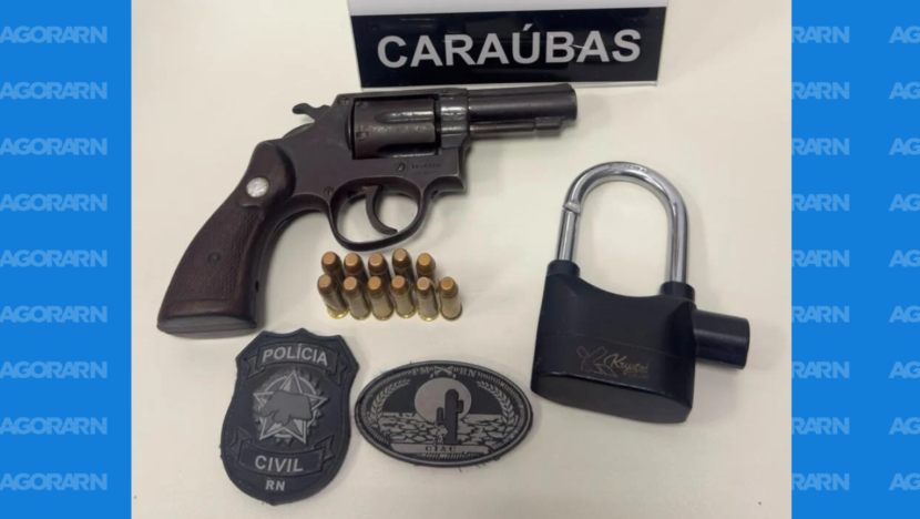 Revólver, munições, cadeado e distintivos da Polícia Civil dispostos sobre uma superfície, com placa de Caraúbas ao fundo