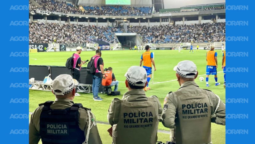 Policiais militares posicionados à beira do campo durante partida de futebol com arquibancadas ocupadas na Arena das Dunas