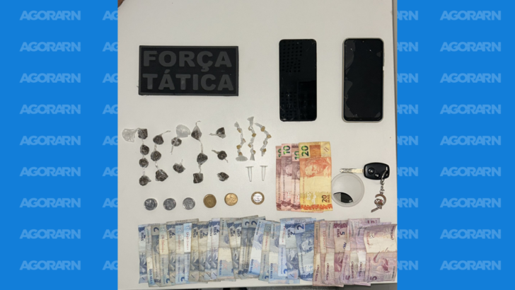 Material apreendido disposto sobre mesa, incluindo porções de substâncias, cédulas e moedas, dois celulares e uma chave de veículo.
