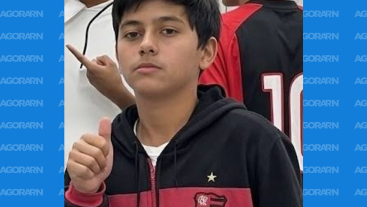 Jovem aparece usando agasalho preto e vermelho e fazendo sinal de positivo com a mão