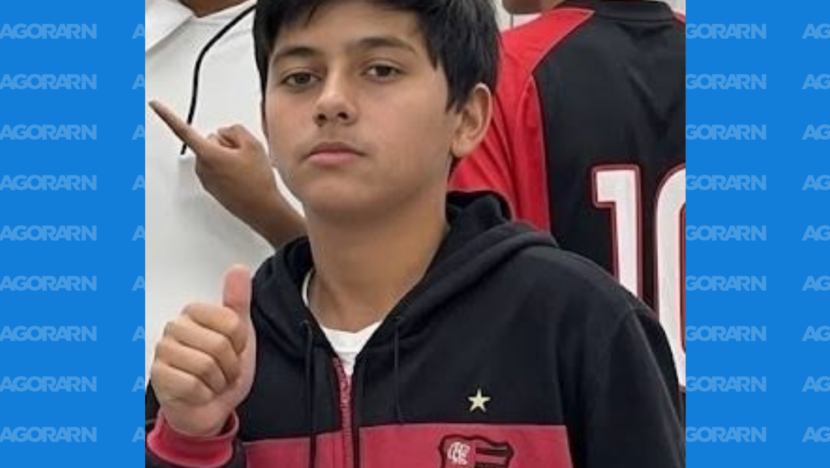 Jovem aparece usando agasalho preto e vermelho e fazendo sinal de positivo com a mão