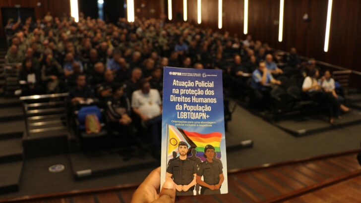 Mão segura cartilha sobre proteção dos direitos humanos da população LGBTQIAPN+ diante de auditório ocupado por agentes de segurança