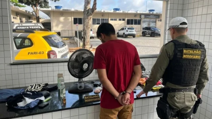 Homem aparece algemado ao lado de policial do Batalhão Rodoviário, com objetos recuperados dispostos sobre um balcão
