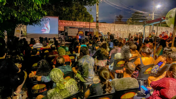 Público sentado em cadeiras do CineSolar assiste a um filme ao ar livre, à noite, ao lado do furgão do projeto