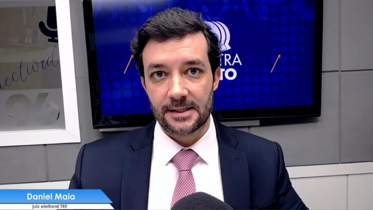 Daniel Maia, de terno escuro e gravata rosa, aparece diante de uma câmera durante entrevista em estúdio