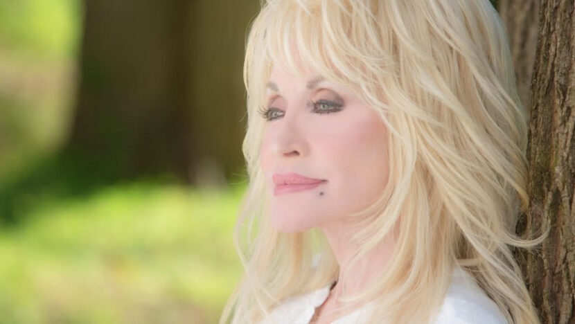 Dolly Parton sorri em retrato ao ar livre, com cabelo loiro volumoso e blusa clara.